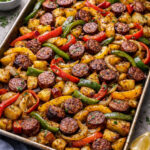 Sheet pan with roasted sausage slices, potatoes, red and yellow peppers, and onions, garnished with herbs.