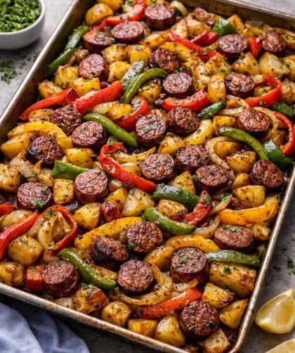 Sheet pan with roasted sausage slices, potatoes, red and yellow peppers, and onions, garnished with herbs.